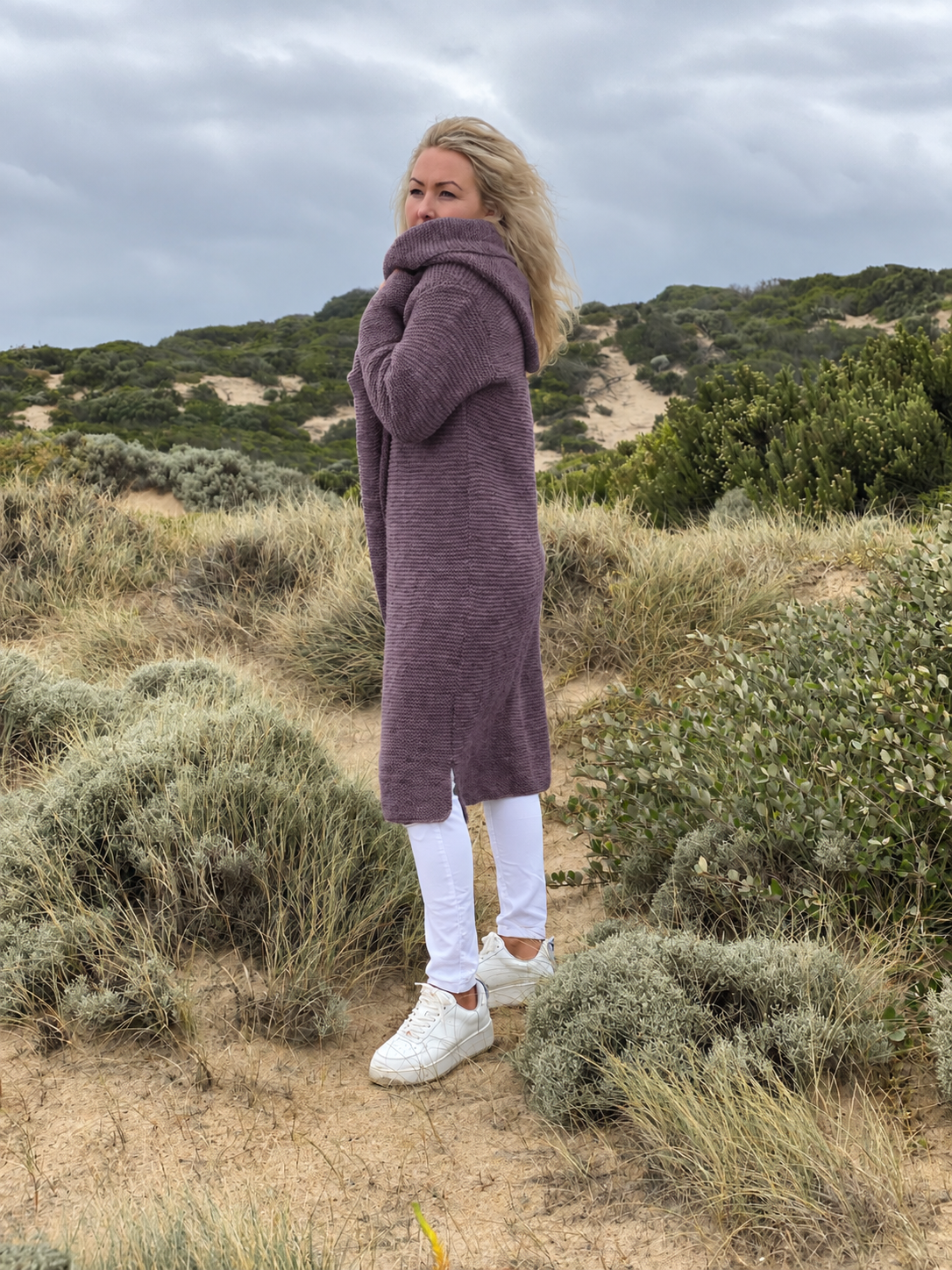 Elna hooded cardigan #133 in grape, a long oversized knit worn with white jeans and trainers by the sea