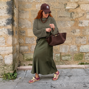 Tora Loose Sweater #141 in eucalyptus, worn over a long green skirt by a stone wall