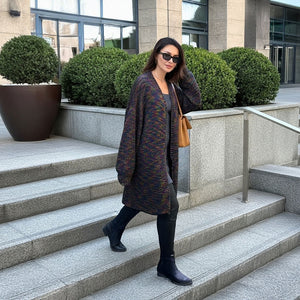 A woman wearing a long, loose dark knit cardigan with subtle multicolor flecks, black pants, and black ankle boots walks down outdoor stone steps. She is carrying a tan shoulder bag and wearing sunglasses.