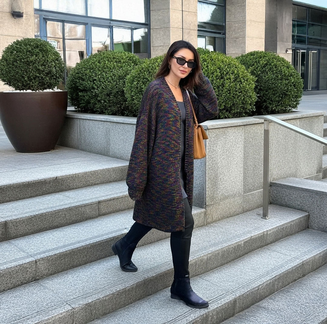 A woman wearing a long, loose dark knit cardigan with subtle multicolor flecks, black pants, and black ankle boots walks down outdoor stone steps. She is carrying a tan shoulder bag and wearing sunglasses.