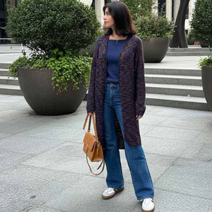 A woman stands outdoors on a paved walkway wearing a long dark purple knit cardigan over a navy top, blue wide-leg jeans, and white sneakers, while carrying a tan handbag.