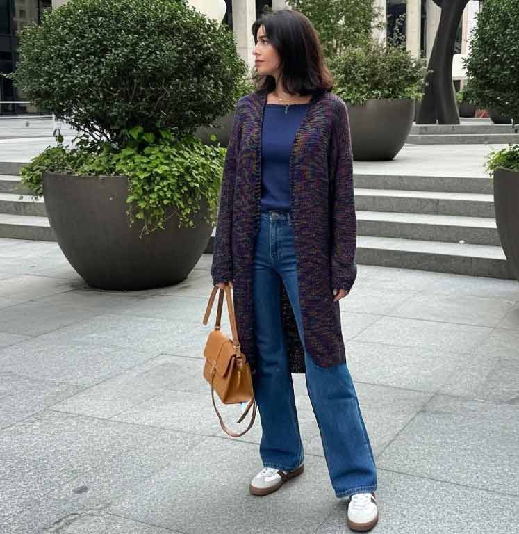 A woman stands outdoors on a paved walkway wearing a long dark purple knit cardigan over a navy top, blue wide-leg jeans, and white sneakers, while carrying a tan handbag.
