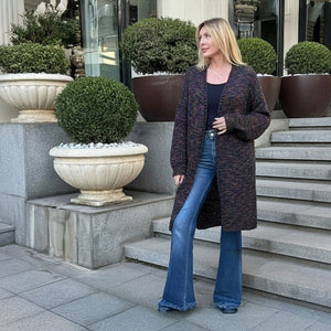 A woman stands on outdoor stone steps wearing a long dark marled cardigan over a black top and blue flared jeans, with large potted shrubs and a building entrance in the background.