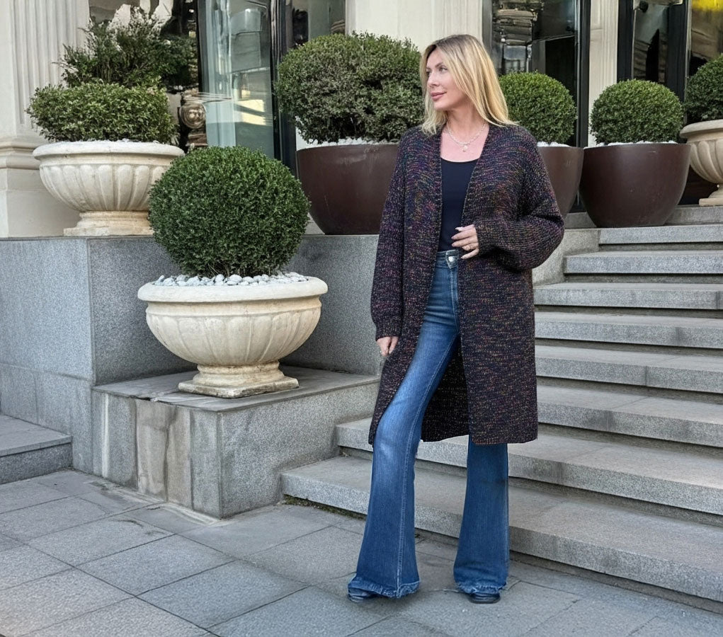A woman stands on outdoor stone steps wearing a long dark marled cardigan over a black top and blue flared jeans, with large potted shrubs and a building entrance in the background.