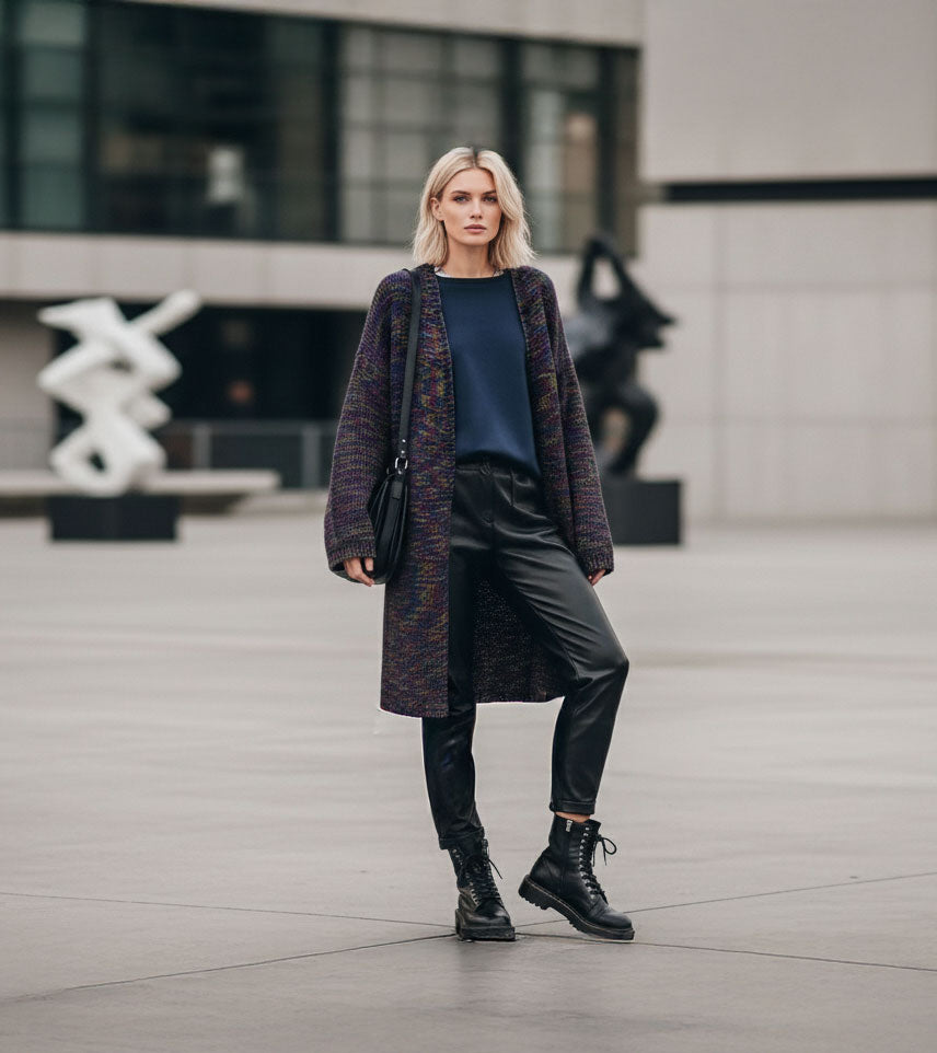 A woman stands in an outdoor urban plaza wearing a long multicolor knit cardigan over a navy top, black leather-look pants, black combat boots, and carrying a black shoulder bag.