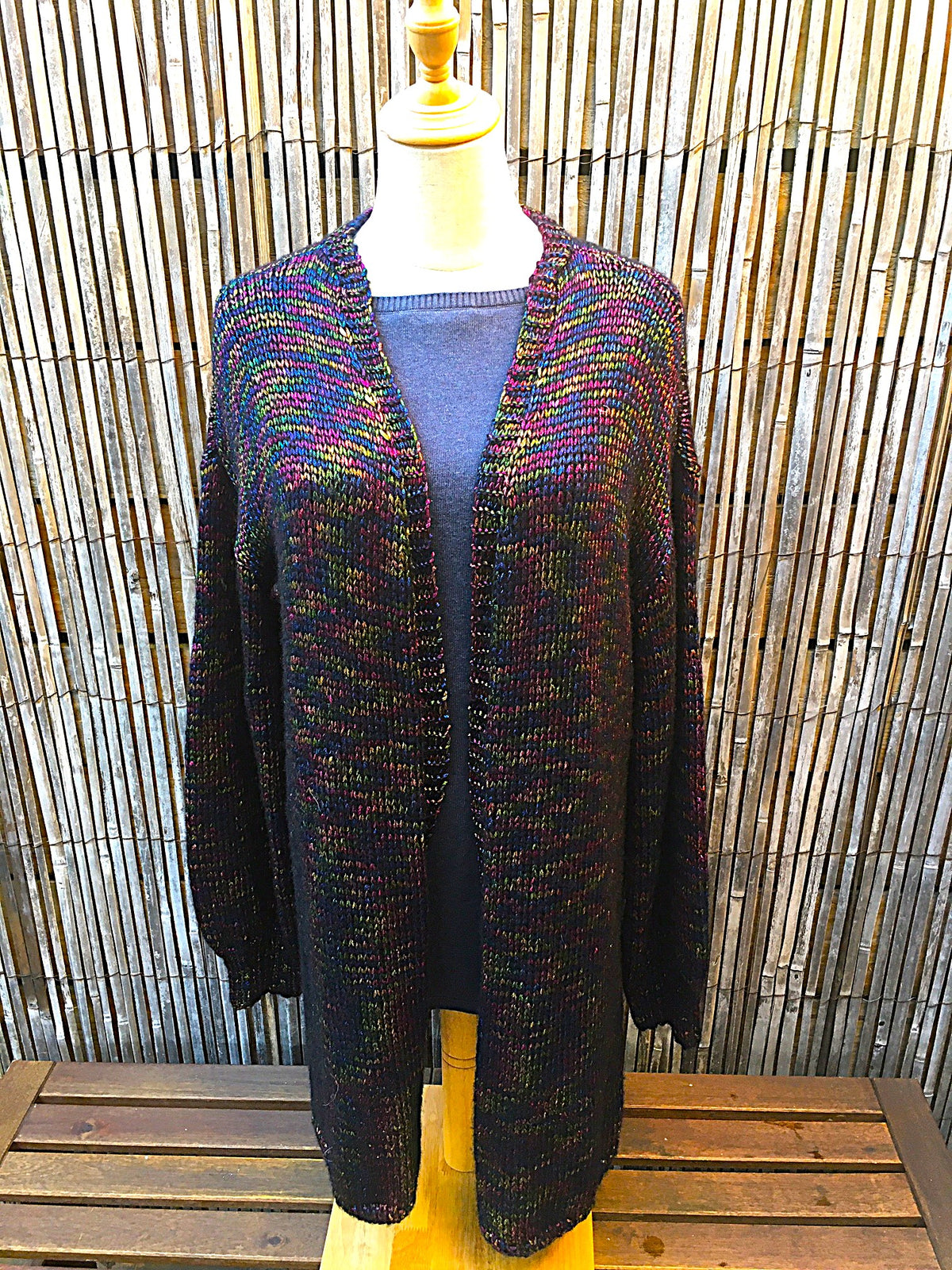 A multicolor knit cardigan sweater displayed on a mannequin torso, featuring a black base with vibrant rainbow flecks, set against a bamboo screen background.