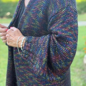 A person outdoors wearing a long, open-front knitted cardigan with a multicolored variegated pattern of blue, green, purple, and orange yarn.