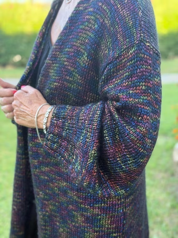 A person outdoors wearing a long, open-front knitted cardigan with a multicolored variegated pattern of blue, green, purple, and orange yarn.