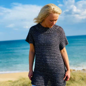 A blonde woman wearing a dark charcoal gray chunky knit tunic top and light blue ripped jeans, standing on a grassy beach dune with the ocean and blue sky in the background.