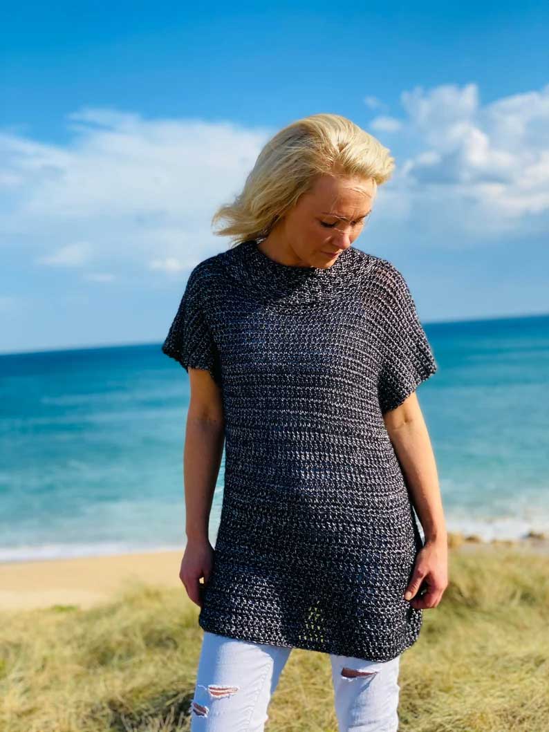 A blonde woman wearing a dark charcoal gray chunky knit tunic top and light blue ripped jeans, standing on a grassy beach dune with the ocean and blue sky in the background.