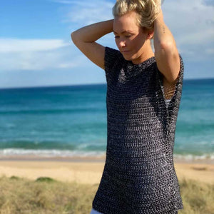 A woman stands on a beach dune wearing a black and white textured crochet tunic top and white pants, with the ocean and blue sky in the background.
