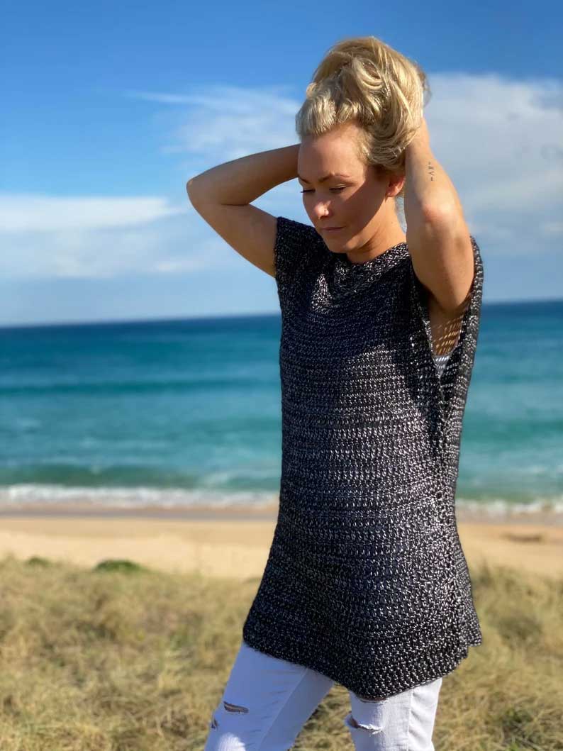 A woman stands on a beach dune wearing a black and white textured crochet tunic top and white pants, with the ocean and blue sky in the background.