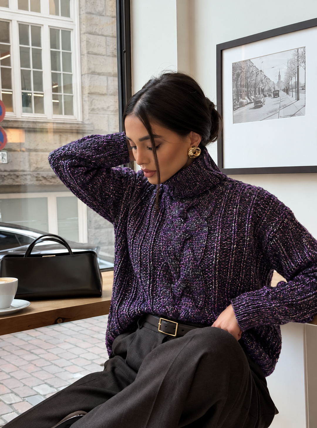 Hedda Cable Roll Neck #150 in amethyst, worn seated in a café with dark trousers