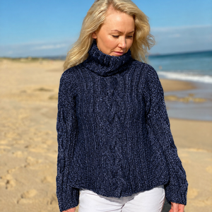 Hedda Cable Roll Neck #150 in marina, an oversized cable jumper worn with white jeans on the beach