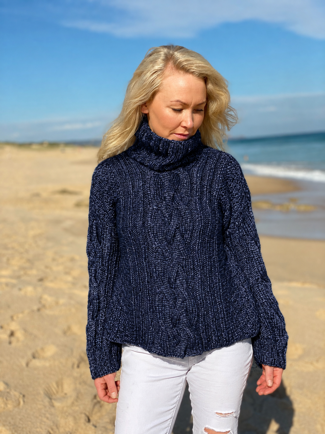 Hedda Cable Roll Neck #150 in marina, an oversized cable jumper worn with white jeans on the beach