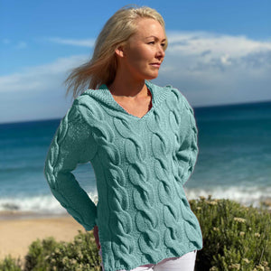 Woman on the beach wearing the Hooded Cable Jumper #172 knitted in Scandi Soft XL Aqua