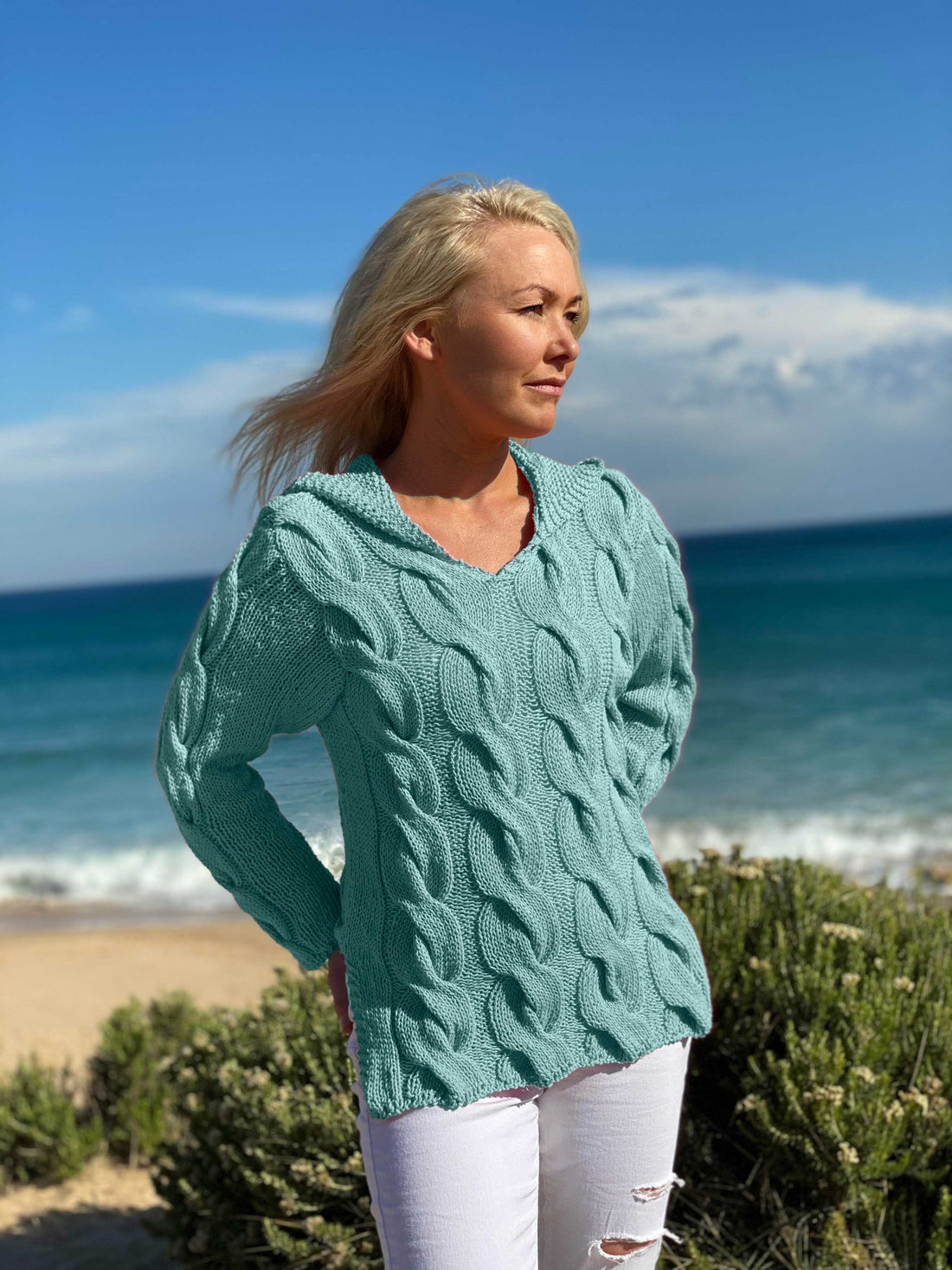 Woman on the beach wearing the Hooded Cable Jumper #172 knitted in Scandi Soft XL Aqua