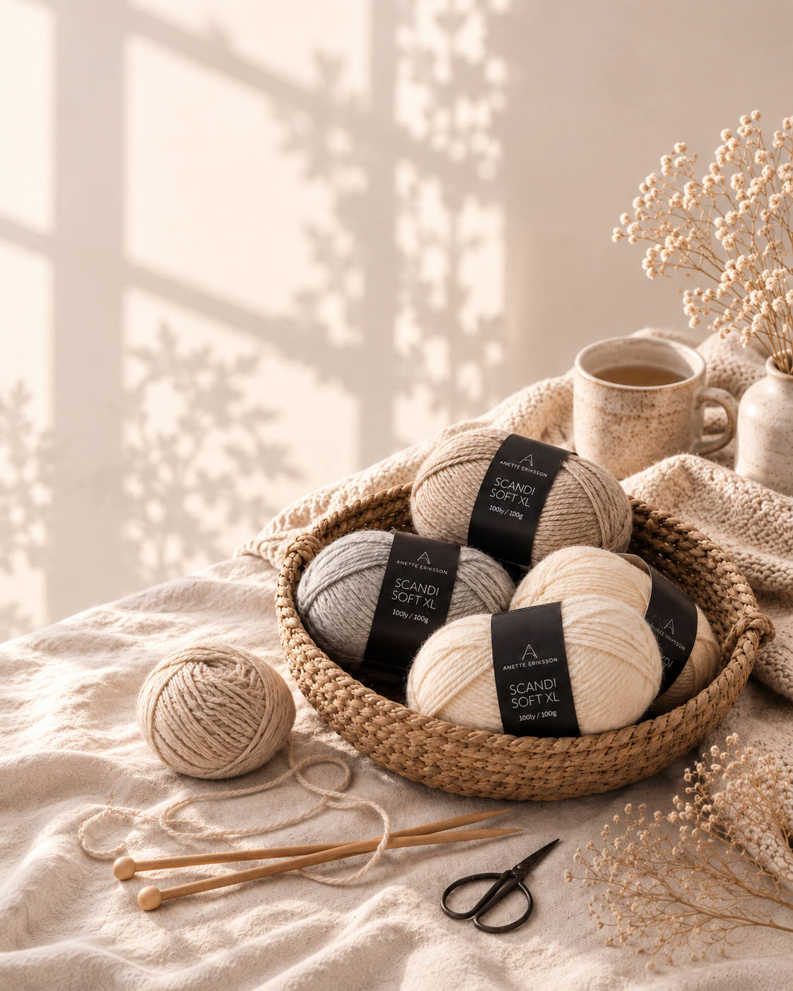 Four balls of yarn in beige, cream, and light gray, each with a black label reading 'SCANDI SOFT', arranged in a woven basket on a beige textile surface, with knitting needles, scissors, a mug, and dried flowers in a softly lit interior scene.