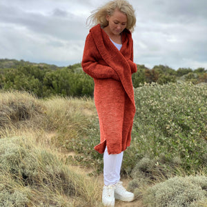 Woman wearing the Elna hooded cardigan #133 in terracotta, a long garter stitch cardigan knitted in chunky Scandi Soft XL, on coastal dunes