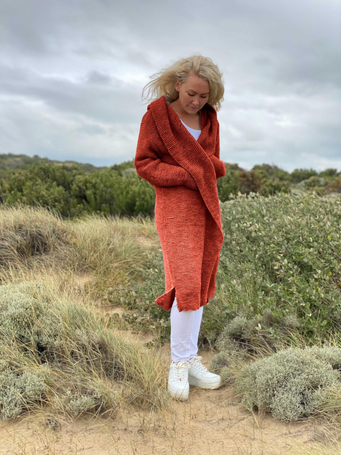 Woman wearing the Elna hooded cardigan #133 in terracotta, a long garter stitch cardigan knitted in chunky Scandi Soft XL, on coastal dunes